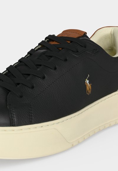 Baskets en cuir noir avec semelle crème, languette marron, lacets noirs et petit logo brodé de joueur de polo sur le côté.