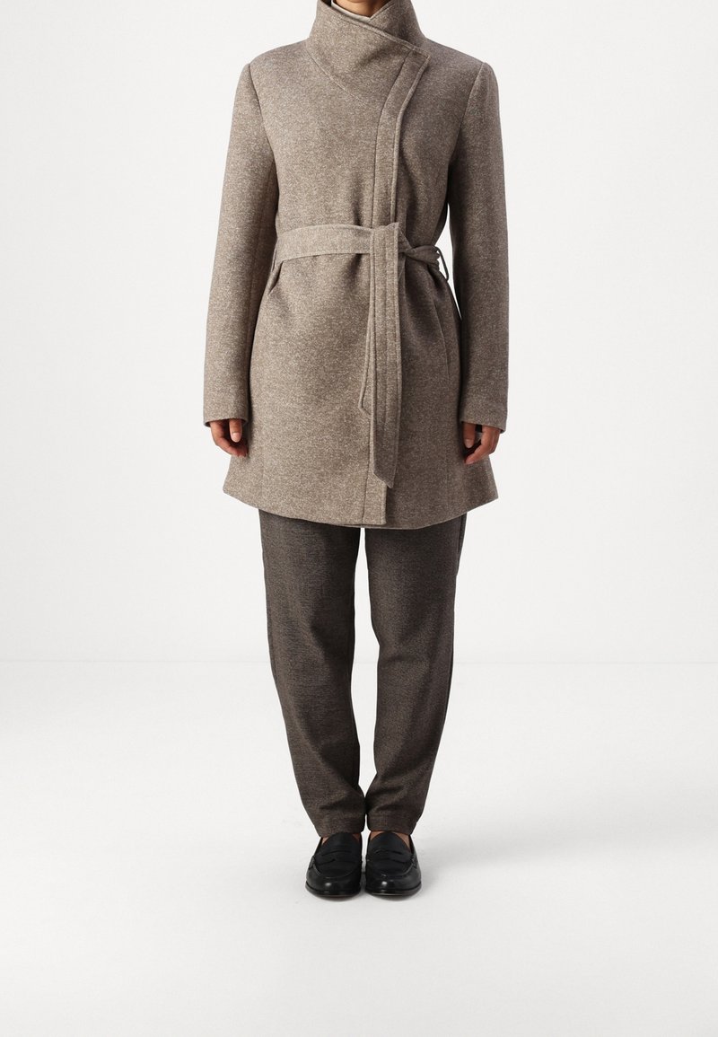 Manteau en mélange de laine marron avec un col haut, une coupe portefeuille et une ceinture à la taille, associé à un pantalon texturé marron foncé et des chaussures noires.