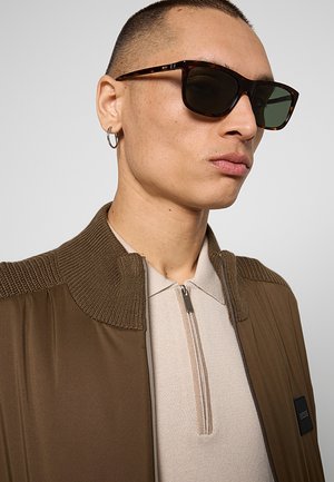 Persona con occhiali da sole tartarugati, piccolo orecchino a cerchio, camicia beige con zip e giacca marrone con collo a maglia e dettaglio sulla spalla.