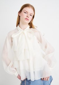 Hvit, lagdelte, gjennomsiktige bluser med volanger, en sløyfe ved halsen, og puffete lange ermer. Myk tekstur, lett stoff.