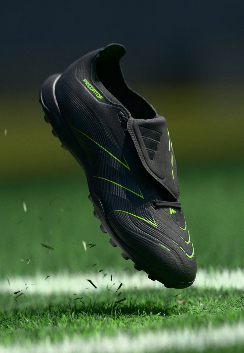 Black soccer shoe with green accents, streamlined design, textured upper, and protruding studs for traction, positioned above grass.