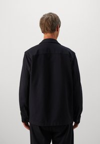 Persona con capelli corti castano chiaro, che indossa una giacca nera a maniche lunghe e pantaloni neri, in piedi di fronte a uno sfondo grigio chiaro uniforme.