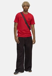 Rød kortærmet skjorte med rund halsudskæring og sort Puma-logo, parret med løse sorte bukser og sort/hvide sneakers.