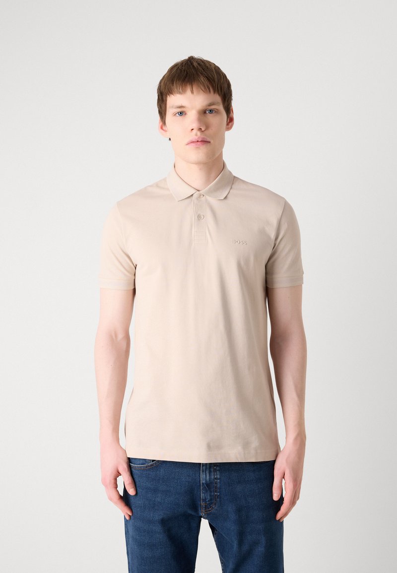 Polo shirt en coton beige avec un col, patte de boutonnage à deux boutons et logo en relief sur la poitrine. Porté avec un jean en denim bleu.