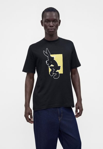 Homme portant un T-shirt noir avec un carré jaune et une silhouette noire de lapin de dessin animé, associé à un jean bleu foncé, debout devant un fond uni.