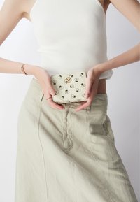 Portefeuille en cuir beige avec des motifs de abeilles noires ; il présente un accent en métal doré et une forme arrondie, tenu entre des mains sur fond neutre.