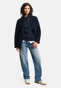 Blazer navy sopra una felpa scura con logo in rilievo, abbinato a jeans blu chiaro e sneakers bianche. Outfit casual con silhouette rilassata.