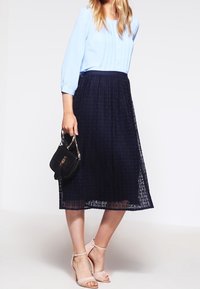 Blusa azzurro chiaro con maniche a tre quarti, abbinata a una gonna midi a trama blu navy scuro. Indossata con sandali beige con cinturini e una borsa nera.