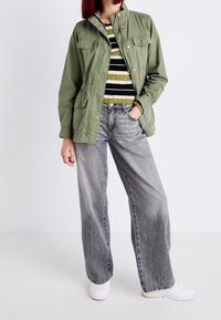 Oliivinvihreä kevyt takki, jossa painonapit, kaksi rintataskua, päällä raidallinen neulepaita ja kulutetut harmaat leveälahkeiset farkut. Valkoiset tennarit.