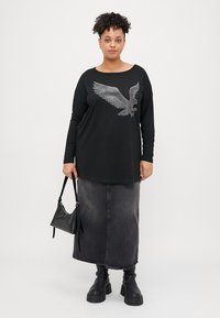 Schwarzes Langarmoberteil mit einem silbernen Adler-Graphic, kombiniert mit einem schwarzen Denim-Rock und klobigen schwarzen Stiefeln. Enthält eine kleine schwarze Handtasche.