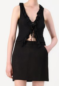 Kvinna iklädd en svart ärmlös volangcrop top med snörning och en svart högmidjad kjol, med handen i fickan mot en enfärgad bakgrund.