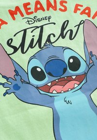 Lys grøn T-shirt med en tegneseriefigur med blå hud, store øjne og et bredt grin. Teksten ovenfor lyder "A MEANS FAIR" og "Disney Stitch."