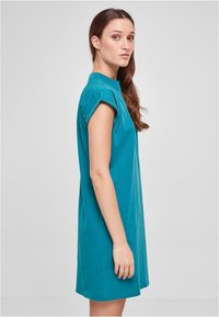 Robe t-shirt en coton turquoise avec col haut et manches courtes, présentant une coupe ample et un ourlet droit, se tenant devant un fond neutre.
