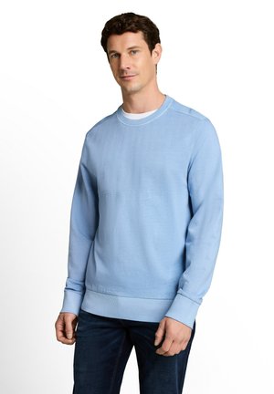 Mann trägt einen hellblauen Pullover mit langen Ärmeln und dunkle Jeans, steht vor einem weißen Hintergrund.