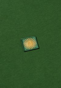 Camiseta de algodón verde con un logo de sol dorado bordado en forma de cuadrado sobre un fondo de tela texturizada.