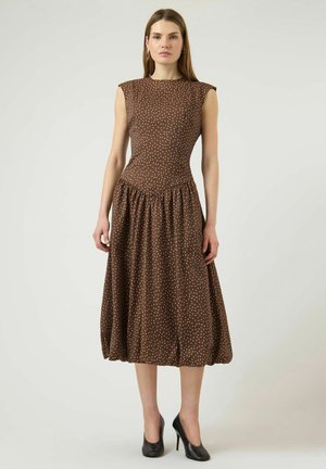 Mujer de pie con un vestido midi sin mangas marrón con lunares blancos y zapatos de tacón negros sobre un fondo claro y sencillo.