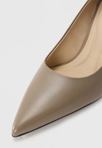 Πέδιλο με μυτερή μύτη από ταπέ ορείχαλκου, με λείο υλικό και χαμηλό άνοιγμα. Το εσωτερικό έχει μπεζ επένδυση και λεία μαύρη σόλα.