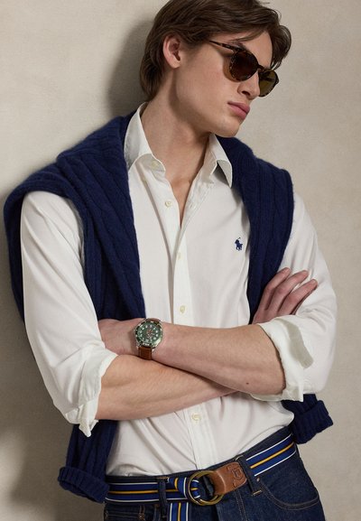 Νεαρός άνδρας που φορά γυαλιά ηλίου με σχέδιο χελώνιας επιφάνειας, λευκό πουκάμισο με λογότυπο, μπλε σκούρο πουλόβερ περασμένο στους ώμους, ριγέ ζώνη και ρολόι με πράσινο καντράν στον καρπό, με σταυρωμένα τα χέρια.