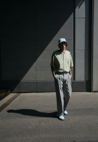 Korte mouwen groene button-up shirt, grijze geplooide broek, lichtblauwe schoenen en een lichtblauwe bucket hat. Staande op gestructureerd asfalt.
