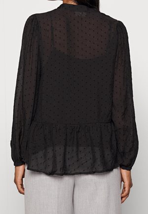 Woman wearing a sheer black long-sleeve blouse with dotted texture and ruffled hem, paired with light gray pants, shown from the back.