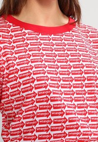 Camiseta roja con un estampado integral de logotipos blancos de "Wrangler" en cajas rojas con forma de flecha, con un cuello redondo rojo y mangas cortas.