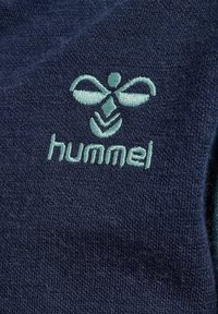 Marinblått tyg med ett strukturerat mönster; har en broderad ljusblå logotyp och ordet "hummel" under logotypen.
