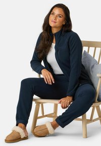 Marine fleece jas en broek, gecombineerd met tan pantoffels met een crèmekleurige bontvoering. Model zit op een houten stoel met kussen.
