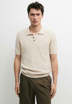 Jeune homme portant un polo tricoté beige à manches courtes avec des boutons foncés et un pantalon vert olive, debout devant un fond blanc uni.