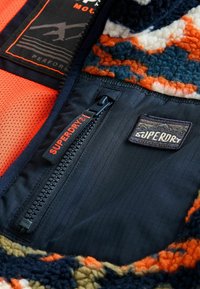 Wnętrze kurtki polarowej z pomarańczową siateczką, ciemnoniebieskim materiałem, teksturowanymi wzorami pomarańczowymi, granatowymi i kremowymi, zamkiem Superdry oraz detalami metki.