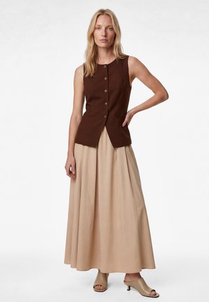 Blonde vrouw draagt een mouwloos bruin vest met knopen, een beige lange rok en beige open hakken, staand met één hand op de heup.