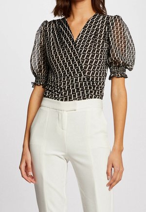 Mujer con blusa de manga abullonada y estampado geométrico en blanco y negro y pantalones blancos de talle alto sobre un fondo liso.