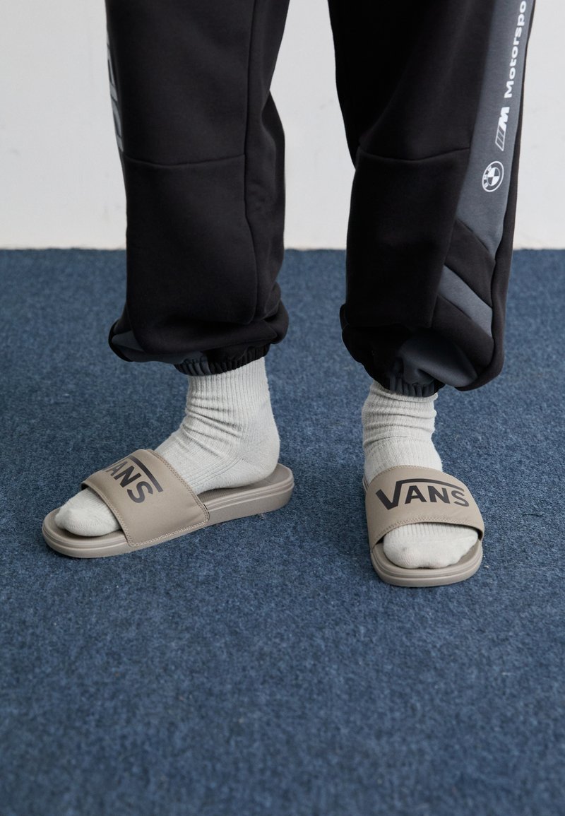 Sandalias deslizantes marrones con una tira ancha que presenta el logo "VANS". Llevadas con calcetines cortos grises claros. Pantalones jogger negros con puños elásticos. Fondo de alfombra azul.