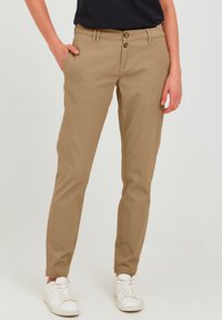 Chinos en coton beige avec une coupe slim, dotés de deux fermetures à bouton, de poches latérales et de revers, associés à des baskets blanches.