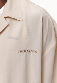 Beige skjorta med krage gjord av mjukt tyg, med subtil brodyr "PEGADOR" och en avslappnad passform med synliga sömdetaljer.