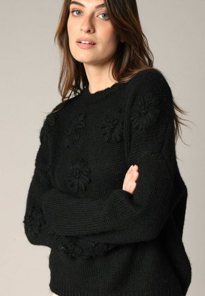 Pull en tricot noir avec des accents floraux, col rond, texture épaisse, et coupe légèrement oversize, avec des poignets et un ourlet côtelés.