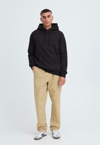 Zwarte hoodie met een voorkinie, gecombineerd met beige broeken en witte sneakers met zwarte en grijze accenten.