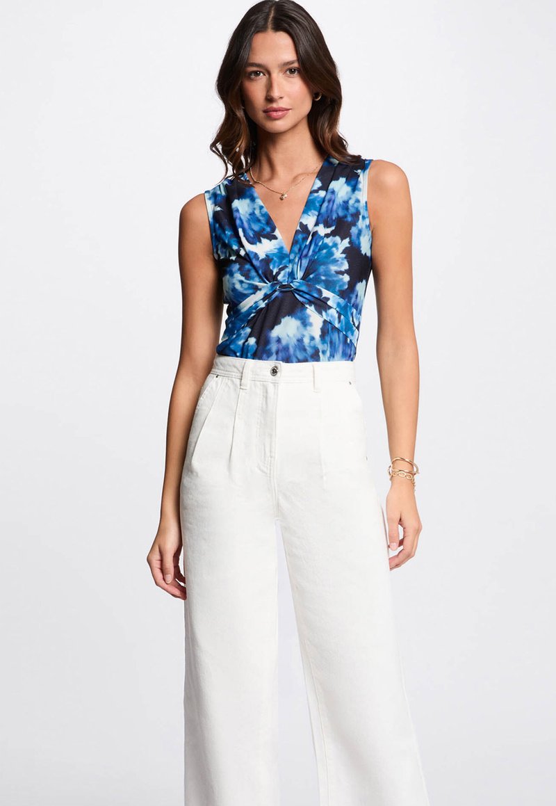 Blau-weißes Batik-Monoshort ohne Ärmel mit V-Ausschnitt und geknotetem Detail an der Taille, kombiniert mit weißen hochgeschnittenen weiten Hosen.