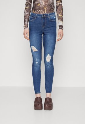 Femme portant un jean skinny bleu déchiré, des sabots marron et un haut à manches longues à motifs, debout devant un fond uni.