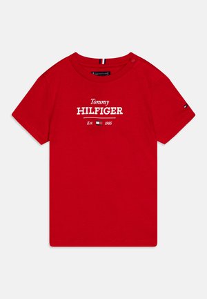 Punainen puuvillapaita, jossa on valkoinen "Tommy HILFIGER" -logo, perustettu 1985, pyöreä kaula-aukko ja lyhyet hihat.