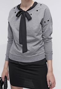 Person som bär en grå sweatshirt med metallöglor och en svart rosett över en svart klänning, hållande en svart handväska.