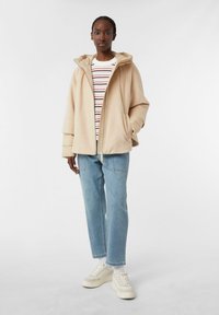 Giacca beige con cappuccio, dalla texture morbida, con zip frontale e grandi tasche, indossata sopra una camicia a righe a maniche lunghe e abbinata a jeans blu chiaro.