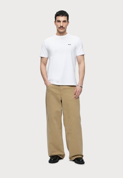 Homme aux cheveux courts et foncés et moustache portant un t-shirt BOSS blanc, un pantalon beige ample et des chaussures noires, debout les mains dans les poches.