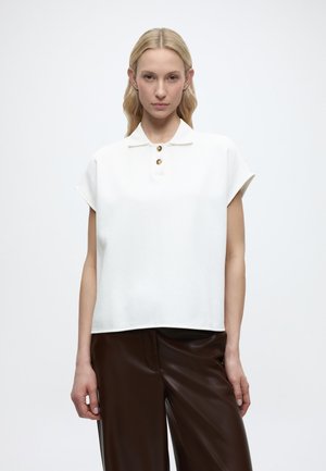 Γυναίκα με μακριά ξανθά μαλλιά φοράει λευκό μπλουζάκι με κοντό μανίκι και γιακά με χρυσά κουμπιά και σκούρο καφέ δερμάτινο παντελόνι, στέκεται μπροστά σε απλό φόντο.