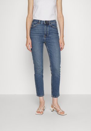 Kobieta nosząca obcisłe niebieskie jeansy z wysokim stanem, w połączeniu z białymi sandałami na obcasie z paskami oraz białą bluzką bez rękawów, stojąca na tle jednolitej ściany.