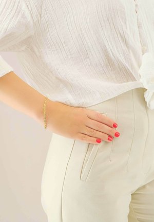 Bracciale a catena d'oro su una mano con unghie rosso acceso. Sono visibili una camicetta bianca con motivo a rilievo e pantaloni a vita alta, su misura, color crema.