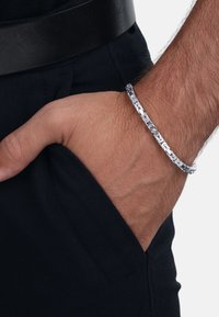 Pulsera de plata con un diseño retorcido, llevada en una mano colocada en un bolsillo de tela oscura, resaltando su superficie reflectante.