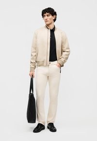 Άνδρας με μπεζ μπουφάν bomber, μαύρο πόλο μπλουζάκι, κρεμ παντελόνι, μαύρα αθλητικά παπούτσια, κρατώντας μαύρη τσάντα tote, στέκεται μπροστά σε λευκό φόντο.