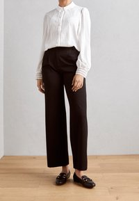 Blouse blanche avec détails en dentelle, pantalon noir à jambes larges et mocassins noirs avec accents de ferrure dorée. Textures lisses et design classique.