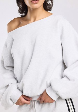 Vrouw draagt oversized lichtgrijze off-shoulder sweatshirt en bijpassende trainingsbroek met zwarte strepen, handen passen de onderkant van de sweatshirt aan.