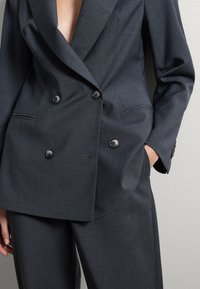 Persona che indossa un blazer doppiopetto grigio scuro e pantaloni abbinati, con una mano in tasca, davanti a uno sfondo parete chiara e semplice.